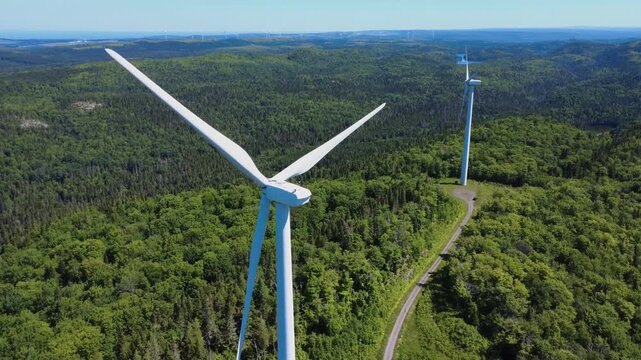 Scenic flight near the head and moving blades of a huge wind turbine atop a verdant mountain range. Saint-Leandre, Quebec, Canada, 2025.