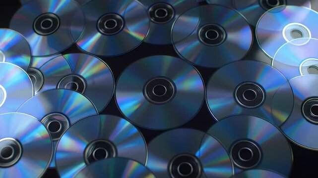 A collection of overlapping compact discs and DVDs reflecting iridescent rainbow colors on a dark surface.