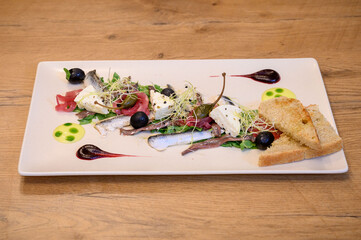 Fine dining seafood starter with sardines, cheese, olives, sprouts, and toast on a white plate with artistic sauces on rustic wooden restaurant table.