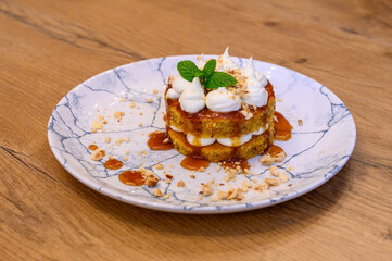 Gourmet dessert with caramel, nuts, sponge cake, and whipped cream on a marble plate, served on a rustic wooden table in fine dining restaurant setting.