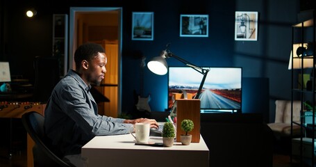 Self employed black man sitting at the desk organizing his schedule, reviewing online data notes. Guy with a coffee cup browsing the internet for research, remote work in a modern loft. Camera A.