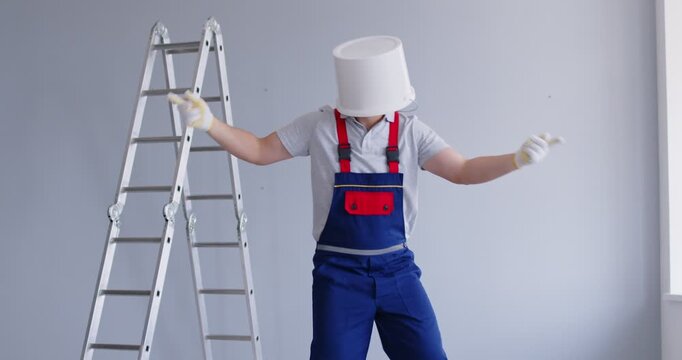 Funny male worker in uniform coveralls, with a paint pucket on his head dancing in a room with grey walls and a step ladder. Home renovations, fun concept