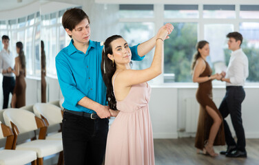 Fototapeta premium Smiling young brunette in light flowing dress practicing sensual bachata with guy in dance class. Social dancing concept..