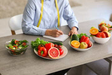 Female nutritionist with healthy food writing on notebook at table in office, closeup