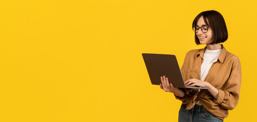 Online education programs. Excited young lady holding and using newest laptop computer, standing over yellow studio background, free space. Happy woman typing on keyboard