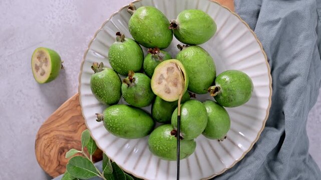 A collection of freshly picked feijoa fruits, some with glistening water droplets, are arranged on a textured white plate, a branch of leaves laying on the side.