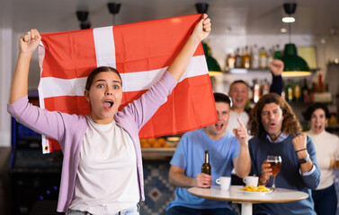 Company of young adult sports fans supporting Danish team with state flag while drinking beer in bar