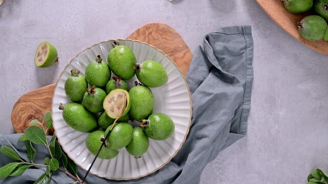 A collection of freshly picked feijoa fruits, some with glistening water droplets, are arranged on a textured white plate, a branch of leaves laying on the side.