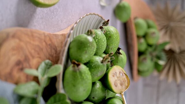 A close-up, high-angle shot of a pile of freshly harvested feijoa fruits, some with glistening water droplets, on a white plate.