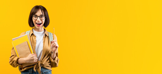 Creative idea. Excited female student pointing pencil up, holding notebooks on yellow studio...