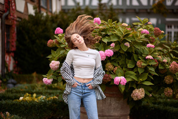 Stylish young woman posing in cozy autumn outfit among blooming hydrangeas in front of traditional half-timbered house in Germany, warm fall colors, outdoor portrait, casual fashion