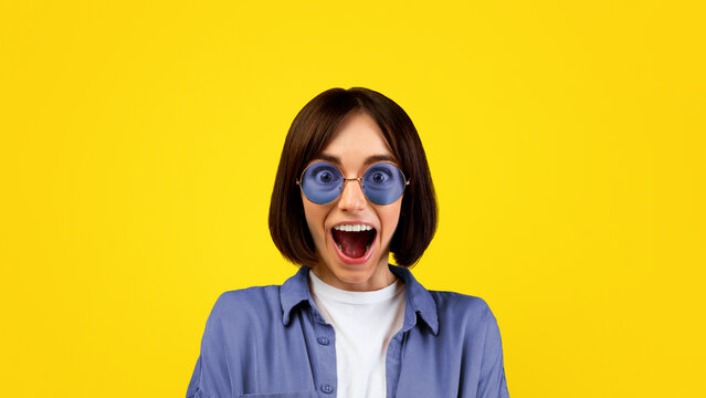 Surprised young woman in colorful sunglasses shouting WOW, opening mouth in excitement over yellow studio background. Happy lady cannot believe shocking news or big sale