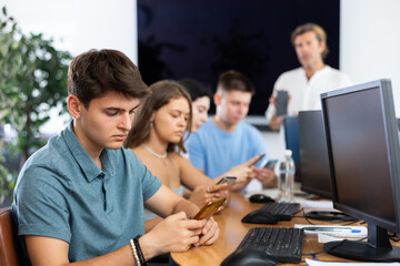 Serious focused young student studying together with coursemates in computer workshop, actively using smartphone to access online learning materials