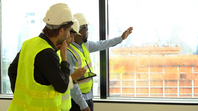 Three colleague architect worker men wear safety vests hard hats looking out window plan talking building site. They are wearing helmet safety vests pointing finger at something building site.Teamwork