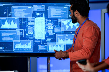 A project manager presents data analytics to his colleagues on a large screen. He is leading a strategy meeting in a high-tech corporate control room or modern office.