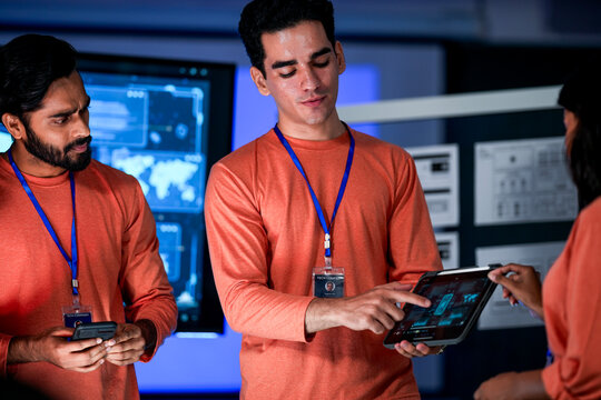 A team of IT specialists in a modern control room works on a project. The diverse group collaborates, reviewing important data on a smartphone and large digital screens.