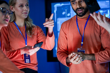 A diverse team of professionals in orange shirts works together in a high tech control room. They are looking at a tablet, discussing a new project or solving a problem.