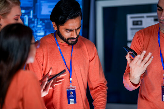 A data analytics team in a command center cross references information from a mobile device with a printed report, while a global data dashboard is displayed on a large screen.