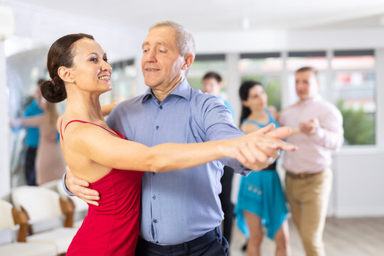 Smiling senior man and young woman dancing slow ballroom dance during group class in choreography studio..