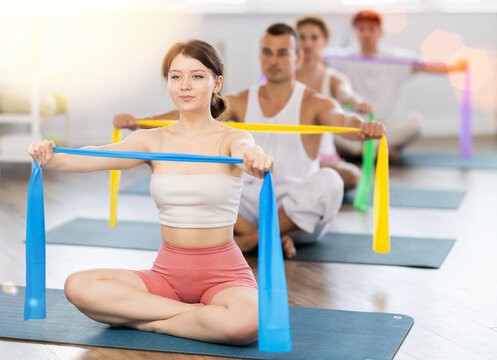 Professional young girl wearing sports clothes using resistance bands during group pilates class sitting on mat in modern fitness room