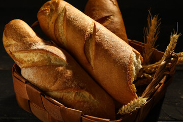 Wicker basket with fresh baguettes and wheat on black wooden table, closeup