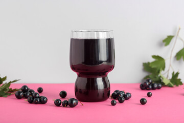 Glass of fresh black currant juice and berries on pink table against grey background