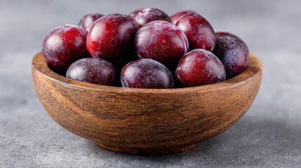 Obraz premium Deep red plums fill a rustic wooden bowl on a gray surface. Close-up view