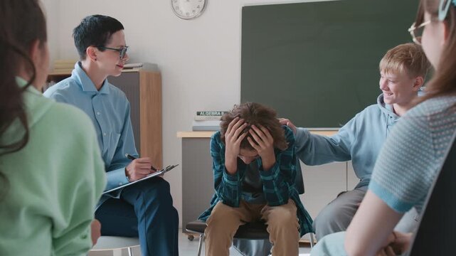 Worried teenage boy receiving support from his friends and school councillor, sitting in circle during group therapy session in classroom