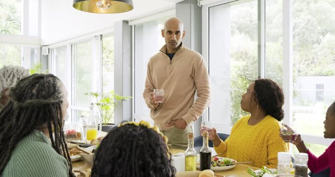 Diverse family father standing at dining table raising glass to toast as everyone clinks glasses
