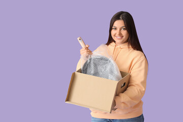 Young woman unpacking frying pan from box on lilac background