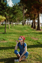 Obraz premium smiling girl sitting on grass wearing sunglasses and denim jumpsuit with red headband enjoying sunny day in park with trees and green lawn around