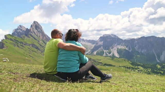 Couple sitting and enjoying peaceful alpine view in the Seceda region of the Dolomites Rear view of couple relaxing on mountain meadow in the Dolomites under blue sky