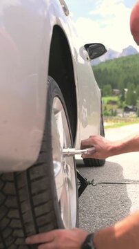 Vertical screen showing man kneeling to fix car tire during roadside emergency in mountain town Man changing flat tire with tools in front of white car