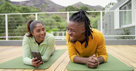 Unlocking phone, diverse couple on rooftop yoga mats swiping clips and laughing with water bottle - Powered by Adobe