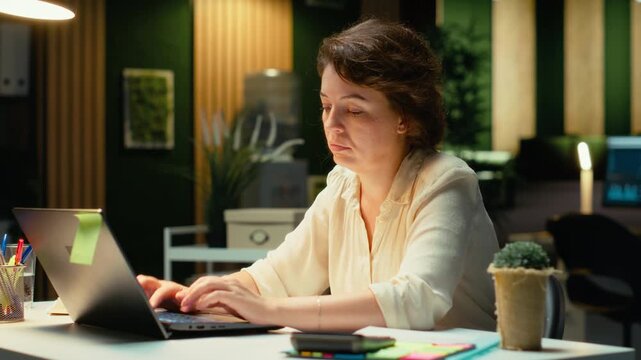Tensed businesswoman stretching her upper back and neck muscles, correcting her chair posture and neck stiffness due to intense office activity. Discomfort and weariness. Camera B.