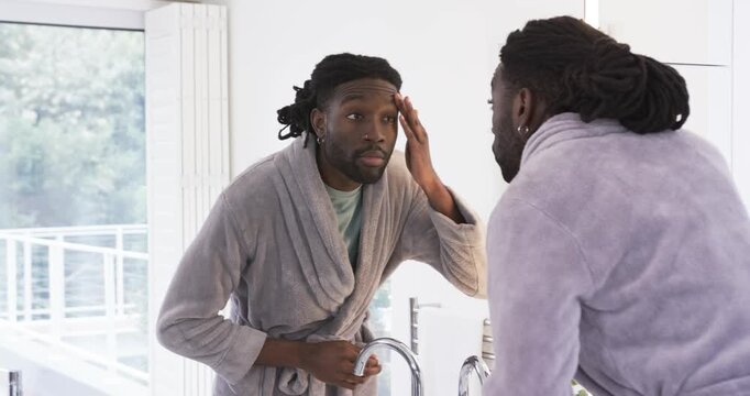 African American man in robe checking mirror spreading cream patting cheeks at sink for skin exam