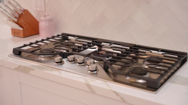 Modern kitchen with built in gas cooktop. Cooktop has five burners and shiny metal knobs. White countertop with soft gray lines. White tile wall with zigzag pattern