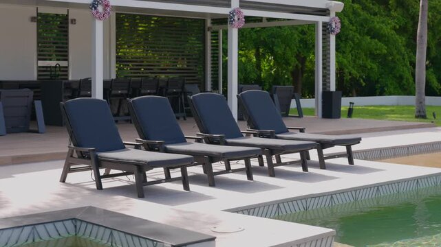 Modern outdoor pool area. Four black lounge chairs on light deck, facing clear swimming pool. Behind them is white pergola with table and chairs under it