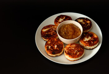 Large round plate with six fresh cheesecakes with raisins and a cup with apple jam on a black background.