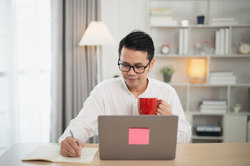 Focused young man working on laptop at home office while holding a red mug, capturing a productive and cozy workspace atmosphere