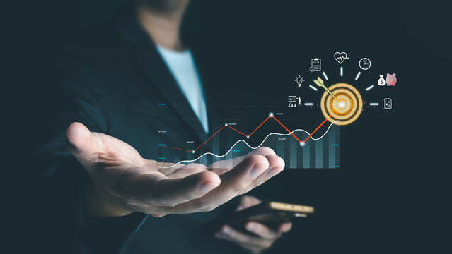 A businessman presents a holographic display of a rising graph and a glowing target with an arrow, symbolizing achieving business goals through modern technology and strategy.