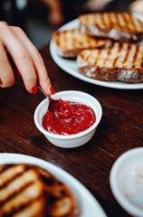 A person with red nails dips a spoon in a white bowl of red jam, with toasted bread on plates on a brown wooden table