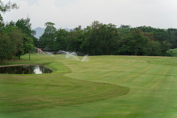 Serene Golf Course Landscape with Sprinklers in Action on the Lush Green Fairway Surrounded by Trees and Reflections in the Still Water Pond