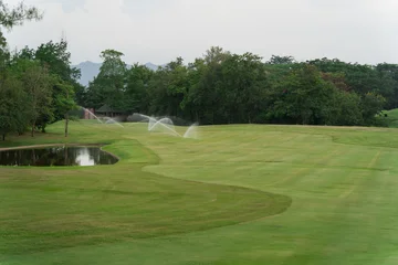 Papier peint photo  Serene Golf Course Landscape with Sprinklers in Action on the Lush Green Fairway Surrounded by Trees and Reflections in the Still Water Pond  © WMSTUDIO