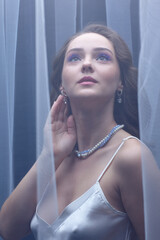 Portrait of a young female covered with veil. Woman is dressed in a peignoir and wears a necklace and earrings.