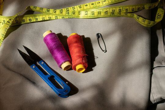 A sewing kit flat lay with thread, scissors, a measuring tape, and a safety pin on a fabric background under dramatic striped lighting.
