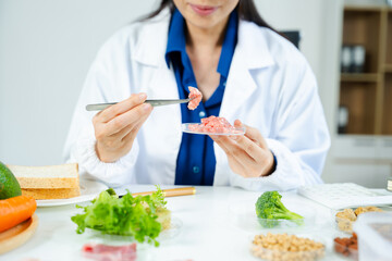 Female scientist in lab coat and goggles examining raw meat with tweezers in food lab. Nutrition, protein, food safety, and research concept.