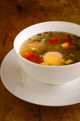 Traditional clear soup with boiled egg, fresh tomatoes, scallions, and vegetables served in a white bowl on rustic wooden table. Homemade Asian comfort food, healthy and warm meal.