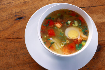 Traditional clear soup with boiled egg, fresh tomatoes, scallions, and vegetables served in a white bowl on rustic wooden table. Homemade Asian comfort food, healthy and warm meal.