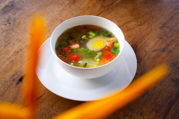 Traditional clear soup with boiled egg, fresh tomatoes, scallions, and vegetables served in a white bowl on rustic wooden table. Homemade Asian comfort food, healthy and warm meal.